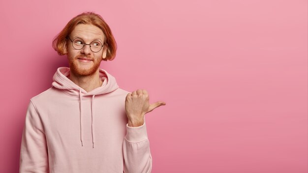 Horizontal shot of ginger bearded Caucasian man points thumb right, wears transparent glasses and hoodie, demonstrates blank space with interest, shows nice promo, isolated over pink wall