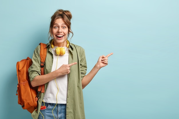 Horizontal shot of glad Caucasian woman points aside with both fore fingers, advertises something aside