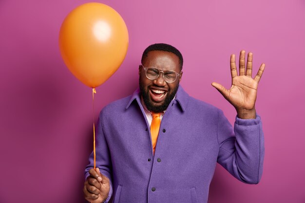 Horizontal shot of glad unshaven man dances carefree, keeps palm raised, laughs positively, holds balloon