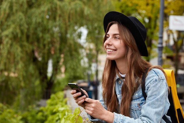 Horizontal shot of glad young teenage girl chats with friend via cell phone, installs app on gadget, wears fashionable black hat