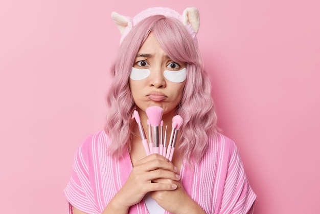 Horizontal shot of gloomy Asian woman with pink hair applies beauty patches under eyes for skin rejuvenation holds cosmetic brushes for applying makeup wears headband poses against rosy wall