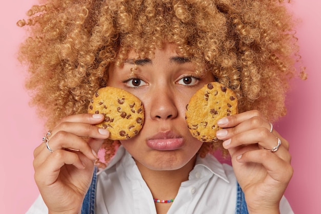 Horizontal shot of gloomy sad woman looks unhappily at camera holds two delicious cookies over face tries to keep healthy diet feels temptation to eat yummy sweet food isolated over pink background