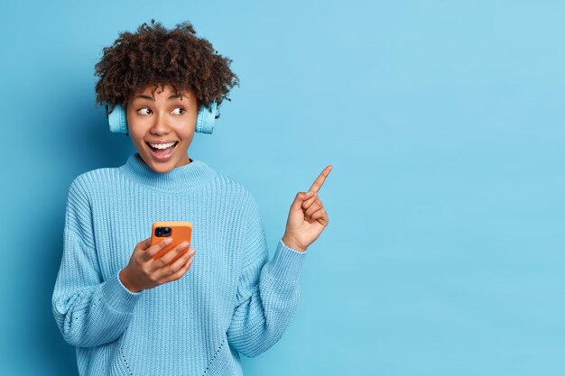 Horizontal shot of good looking Afro American woman feels very happy holds modern smartphone in hand wears stereo headphones points aside on blank space over blue background. Leisure concept