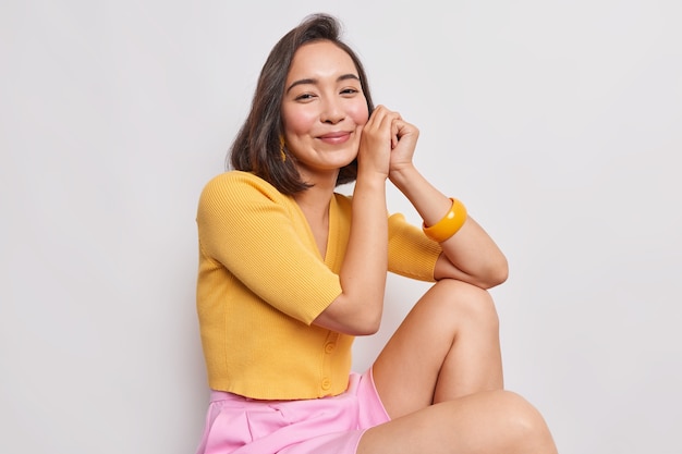 Horizontal shot of good looking Asian woman keeps hands near face smiles pleasantly leans on knee wears yellow jumper isolated over white wall