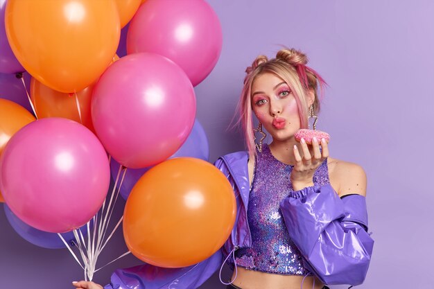 horizontal shot of good looking blonde woman keeps lips folded holds glazed doughnut dressed in stylish clothes has bright makeup holds inflated colorful balloons 