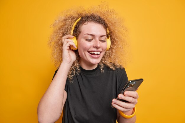 Horizontal shot of good looking young woman with curly bushy hair smiles broadly looks at smartphone display listens audio track via headphones dressed casually isolated over yellow wall