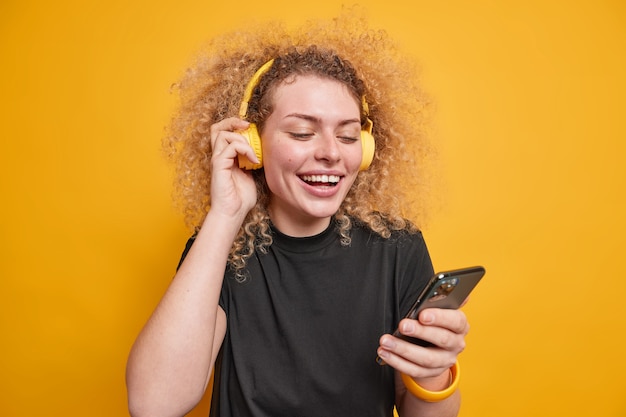 Horizontal shot of good looking young woman with curly bushy hair smiles broadly looks at smartphone display listens audio track via headphones dressed casually isolated over yellow wall