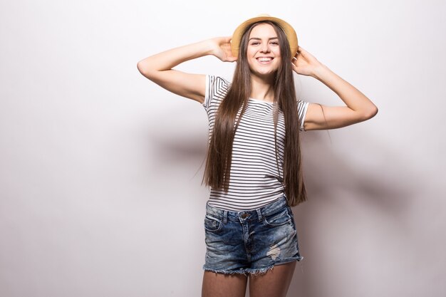 Horizontal shot of gorgeous female model keeps hand on straw hat, wears white top with bare shoulders, looks with confident expression, isolated over white wall