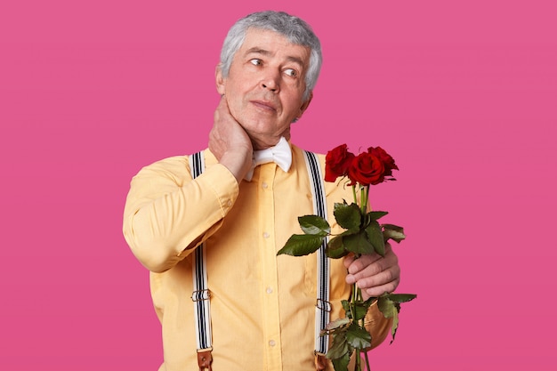 Horizontal shot of grey haired elderly man in formal fashionable clothes, keeps hand on neck, has pain, looks aside, holds red roses