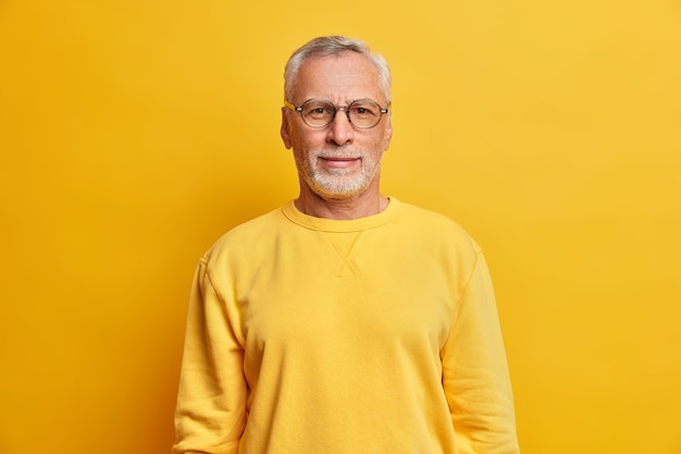 Horizontal shot of grey haired man with wrinkles wears glasses and casual yellow jumper looks directly at front has satisfied expression poses indoor