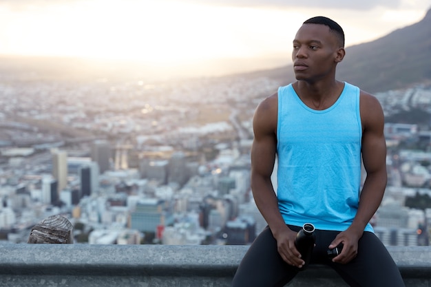 Horizontal shot of handsome black man in casual outfit, holds bottle of water, being deep in thoughts, rests over city with daylight, clear sky with copy space for your promotion.