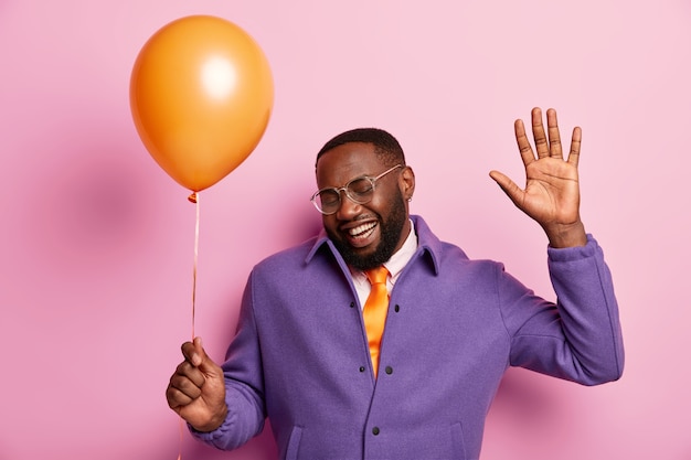 Horizontal shot of handsome black man with thick bristle, dances to music, has fun on party holds air balloon