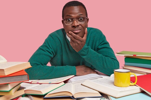 Горизонтальный снимок: красивый черный молодой мужчина держит подбородок, смотрит с любопытством, ищет полезную информацию в книгах, одет в зеленый джемпер.