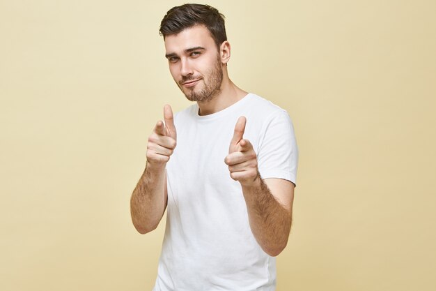 Horizontal shot of handsome charismatic young man with black hair and stubble posing isolated pointing fore fingers, having confident flirty facial expression, choosing you. Body language