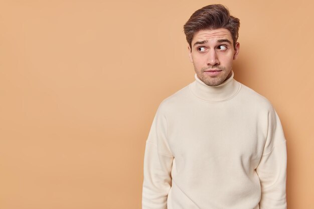 Horizontal shot of handsome dark haired man looks with curious expression at something interesting feels suspicious wears casual turtleneck isolated over beige wall with copy space area for text