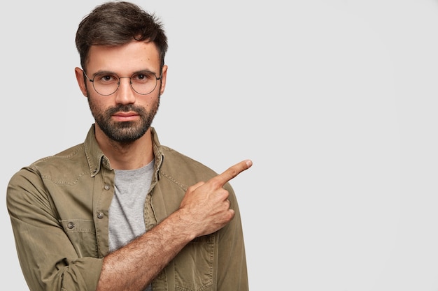 Horizontal shot of handsome unshaven young male with dark stubble, strict look, has serious expression, dressed in fashionable shirt, points right aside