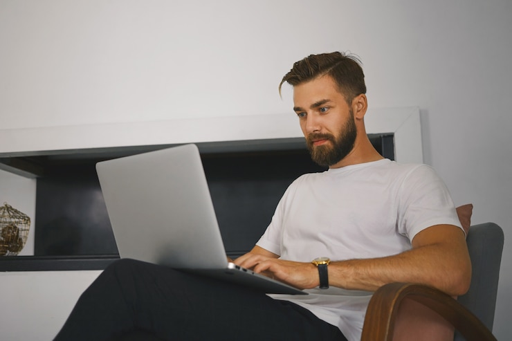 Free Photo horizontal shot of handsome young male freelancer with thick beard sitting in armchair with generic laptop computer, working remotely from home. people, gadgets, technology and communication concept