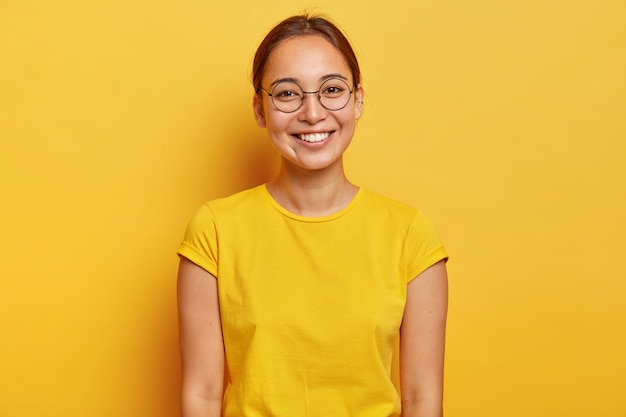 Horizontal shot of happy Asian female student wears big round spectacles, yellow casual wear, smiles gently , satisfied after successful day at university, dressed in summer yellow t shirt