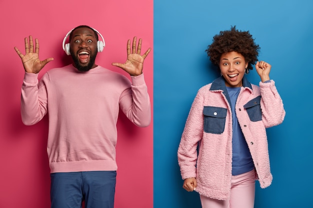 Horizontal shot of happy carefree amused woman and man dance and enjoy music, dressed in casual wear. Glad bearded guy listens awesome song in headphones, raises palms, Afro female keeps hands up