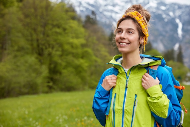 Horizontal shot of happy carefree young woman strolls outside against mountain landscape, enjoys spending free time on meadow