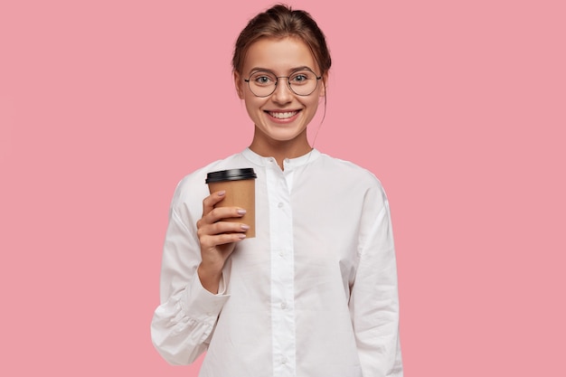 Горизонтальный снимок счастливой кавказской девушки в белой рубашке, несущей бумажный стаканчик с кофе, предлагаю вам выпить