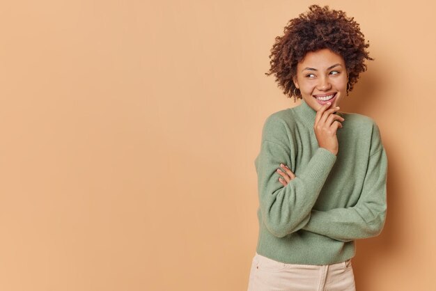 Horizontal shot of happy curly haired woman laughs positively smiles broadly keeps hand on chin wears casual comfortable jumper poses against brown background blank space away for your promo