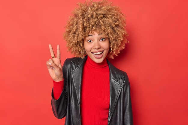 Horizontal shot of happy curly haired woman makes v sign shows peace gesture has glad expression wears turtleneck and leather jacket isolated over vivid red background. Body language concept