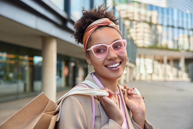 행복한 곱슬머리 여성의 수평 샷은 세련된 핑크색 선글라스 머리띠를 착용하고 재킷은 종이 쇼핑백을 나른다