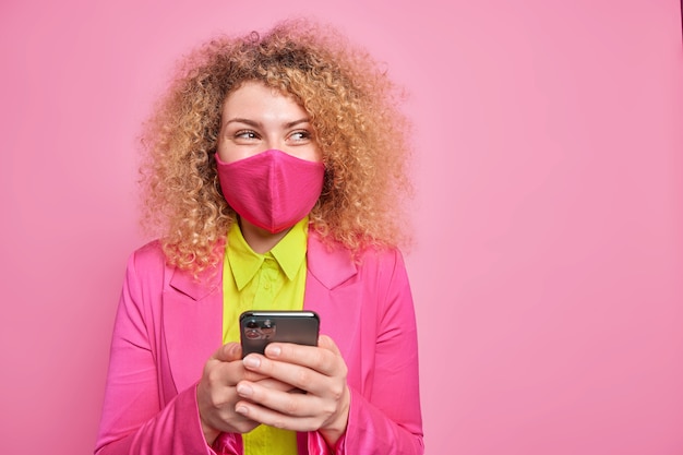 Горизонтальный снимок счастливой кудрявой молодой женщины в защитной маске, задумчиво смотрит в сторону, носит защитную маску для лица и формальная одежда позирует на розовой стене с зоной для копирования