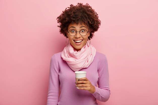 Horizontal shot of happy dark skinned woman with Afro hairdo, holds paper cup with coffee, wears purple jumper and silk scarf