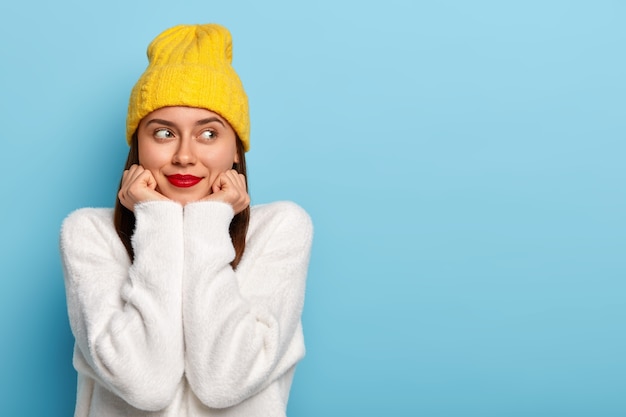 Free photo horizontal shot of happy dreamy european woman wears minimal makeup, red lipstick, looks aside, dressed in yellow hat and white sweater, poses against blue background, being fascinated and pleased