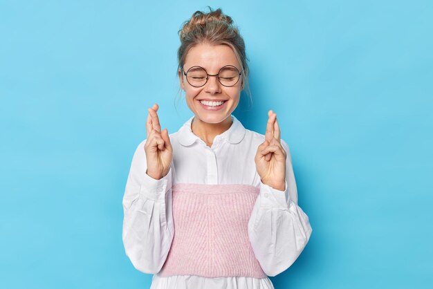 Horizontal shot of happy elegant woman in formal shirt keeps eyes closed crosses finger believes in good luck isolated over blue background. Big hope for dreams come true. Body language concept