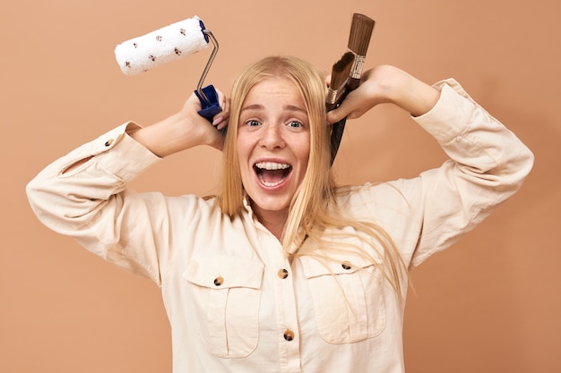 Free photo horizontal shot of happy emotional young blonde female in stylish shirt holding special tools while doing repair in her apartment, excited about renovation