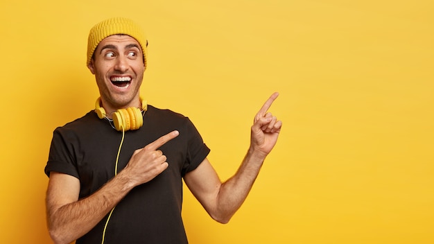 Horizontal shot of happy European man points aside with two index fingers, dressed in stylish black and yellow wear, wears modern headphones around neck for listening song