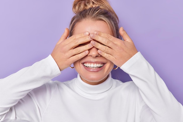 Foto gratuita l'inquadratura orizzontale di una donna europea felice copre gli occhi con le mani attende sorrisi a sorpresa indossa ampiamente un dolcevita bianco casual isolato su sfondo viola attende il miracolo. concetto cieco