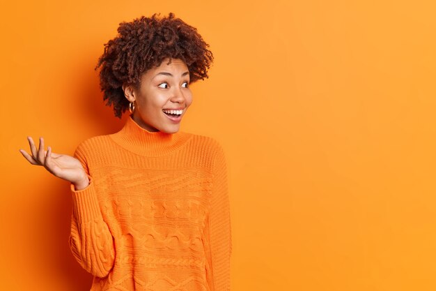 Горизонтальный снимок счастливой возбужденной женщины, поднимающей ладонь, замечающей что-то неожиданное, и удивительной улыбки, положительно одетой в повседневный джемпер, изолированного над ярко-оранжевой стеной