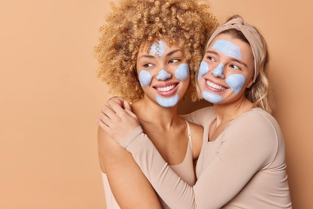 Free photo horizontal shot of happy friendly young women embrace and smile positively apply clay masks on face take care of skin complexion isolated over brown backgound blank copy space for your text