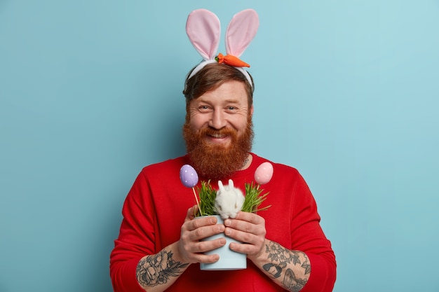 Horizontal shot of happy ginger hipster guy expresses positive emotions, wears bunny ears, has tattoo, holds pot with small rabbit and two decorated eggs, symbols of Easter. Holiday concept.