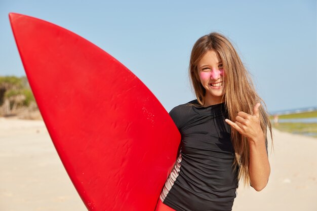 幸せな女の子の水平方向のショットは、サーフィンに適した気象条件を楽しんだり、シャカを作ったり、ゆるいジェスチャーをしたりします