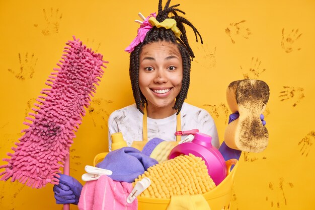 Horizontal shot of happy housekeeper smiles pleasantly being in good mood after finishing cleaning apartment poses near laundry basket isolated over yellow wall