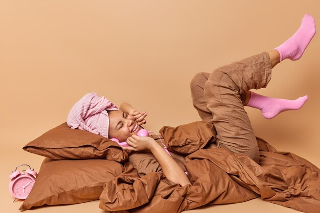 행복한 게으른 여성이 오랫동안 침대에 누워 있는 수평 사진에는 갈색 배경에 대해 침실에서 편안한 잠옷을 입은 복고 핸드셋이 귀 근처에 유지됩니다.