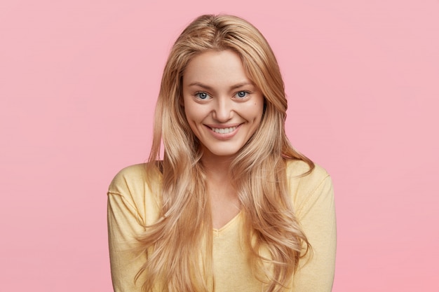 Horizontal shot of happy pleased female model with pleasant smile has long straight hair, wears yellow sweater, glad to recieve good job position, poses against pink wall. Positive emotions