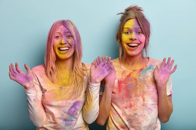 Horizontal shot of happy positive young women raise palms, stand next to each other over blue space