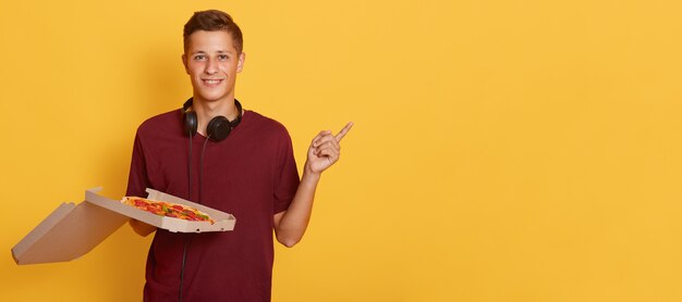 Horizontal shot of happy teenager looking directly  making gesture, showing direction with forefinger, holding box with pizza