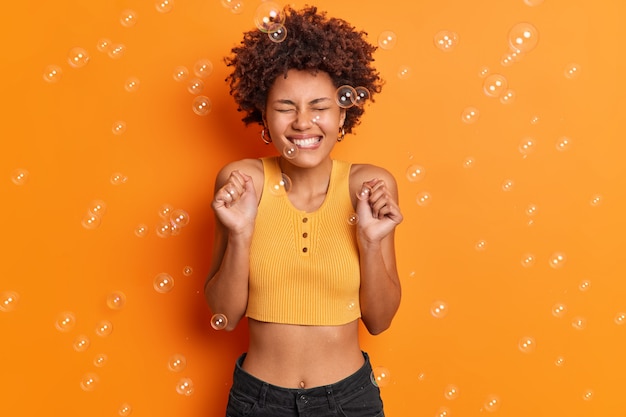 Horizontal shot of happy triumphing woman with curly Afro hair bites lips happy to get approval or hear excellent news wears cropped top and jeans poses against orange wall soap bubbles