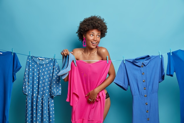 Free photo horizontal shot of happy undressed woman hides behind pink dress hanging on rope, poses near different clothes items, holds shoes in blue color, dresses for job interview, wants to look awesome