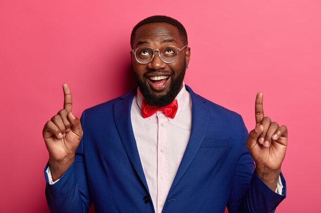 Horizontal shot of happy unshaven Afro American office worker points at copy space above, shows something on blank wall, dressed elegantly