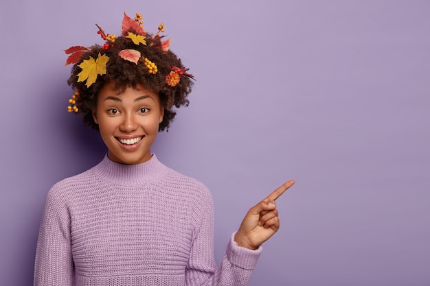 Horizontal shot of happy woman helps to pick up best choice, points fore finger on blank space, smiles pleasantly, wears warm sweater, has curly hairstyle with autumn foliage