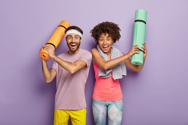 Colpo orizzontale di donna e uomo felici divertirsi dopo l'aerobica, alzare le mani con karemat piegati, vestiti con abbigliamento sportivo, godersi il tempo libero per lo sport, isolato sul muro viola. coppia diversificata