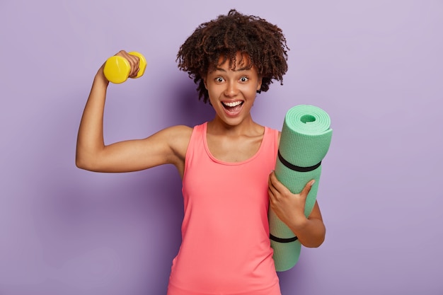 Horizontal shot of happy woman with Afro hairstyle, lifts weight for having biceps, carries rolled up fitness mat, wears pink vest, looks happily, models against purple wall. Sport, motivation